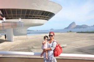 A young woman with a baby in her arms poses in front of a courtyard with a modern looking building and the bay in the distance.