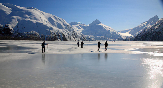 Hikers with trekking poles cross the surface of a frozen lake with snow-capped mountains in the distance.
