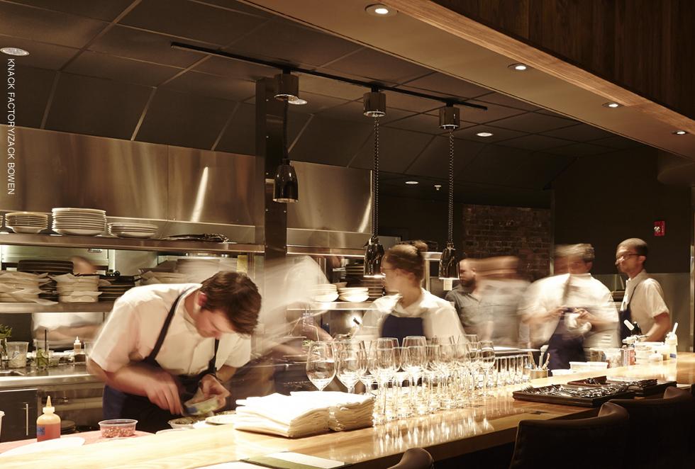 Chefs work in an open kitchen behind a bar with rows of glassware.