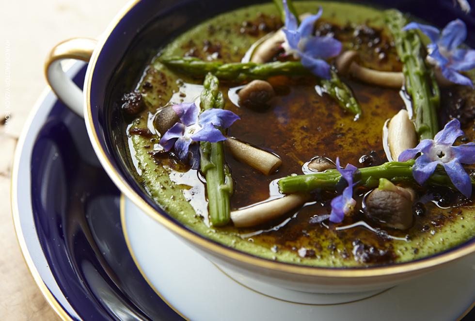 A bowl of soup with fresh asparagus and mushrooms garnished with purple blossoms and served in an elegant yet hearty looking dish.