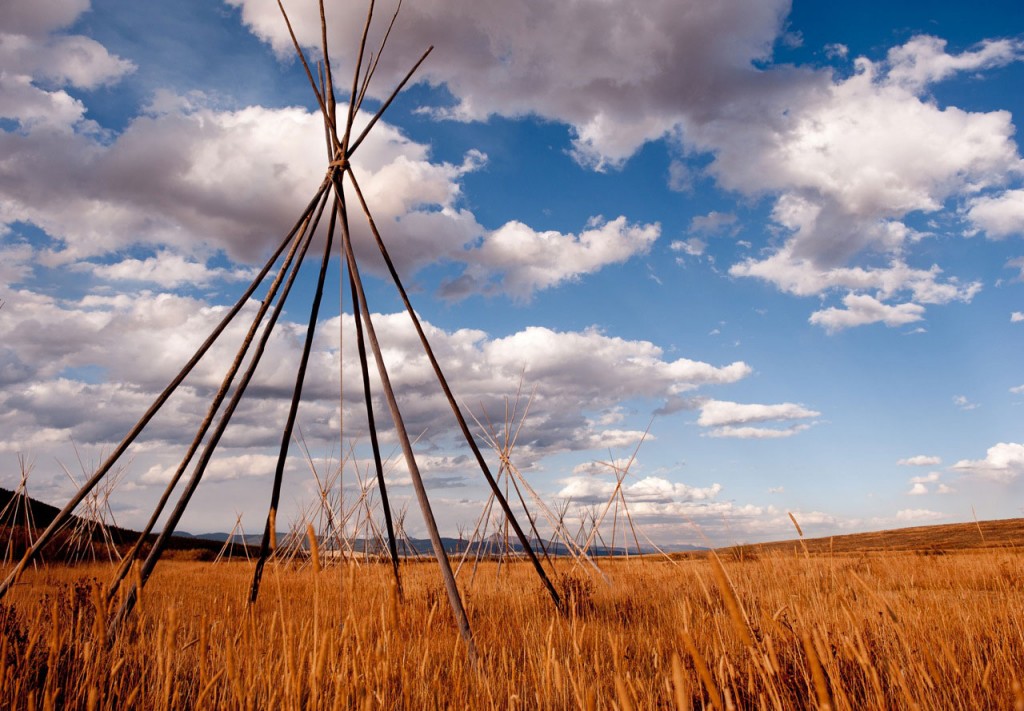 Tipi poles on a golden grassy plain with a dramatic cloudy sky.