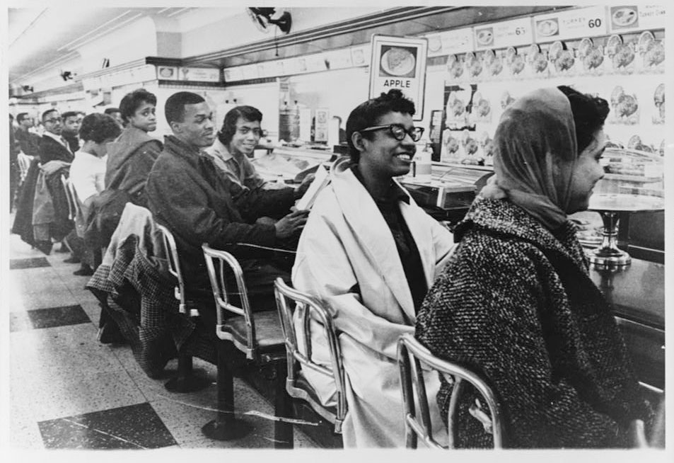 A black and white photograph of a group of college-aged African Americans seated at a lunch counter.