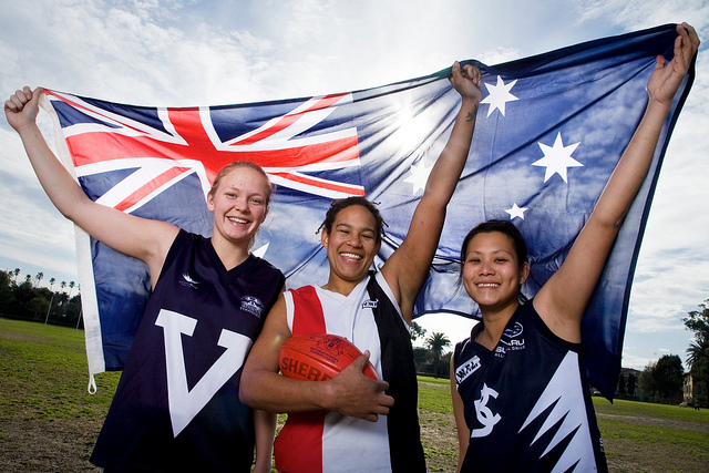 Three girls, holding a football and wearing football jerseys, holding up an Australian flag.