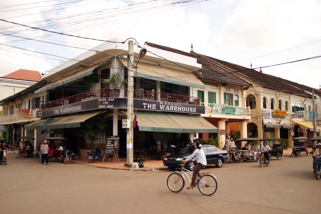 A woman on a bicycle pedals through an intersection in front of a building with banner lettering reading The Warehouse.