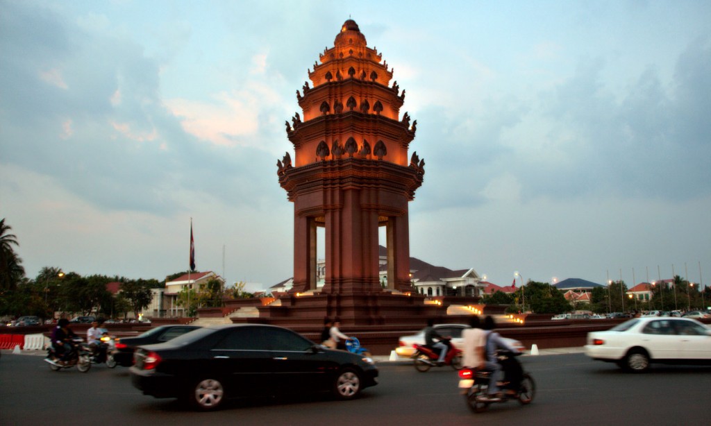 A lit, temple-like structure reminiscent of a lotus blossom sits in the center of a roundabout filled with cars and motorcyclists.