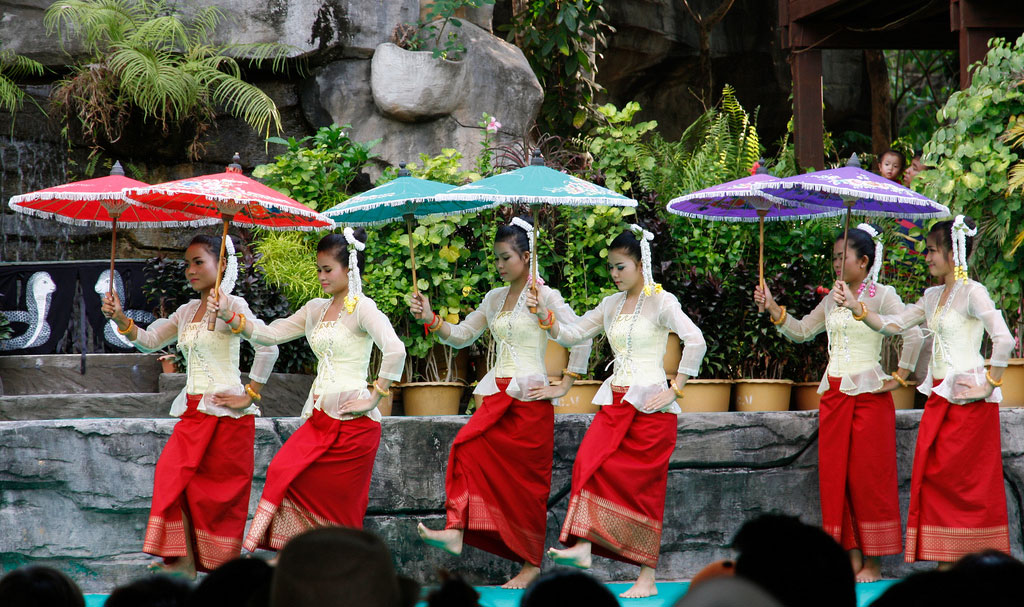 Dancers in matching white tops and red skirts hold colorful parasols as they perform.
