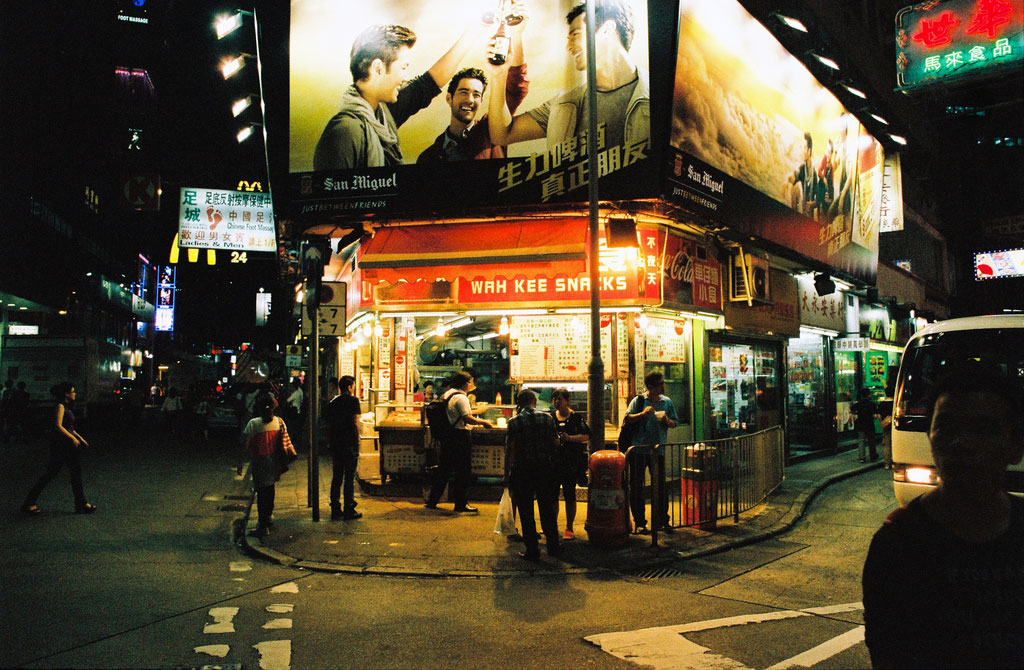 A bus and pedestrians pass by a snack shop on a street corner.