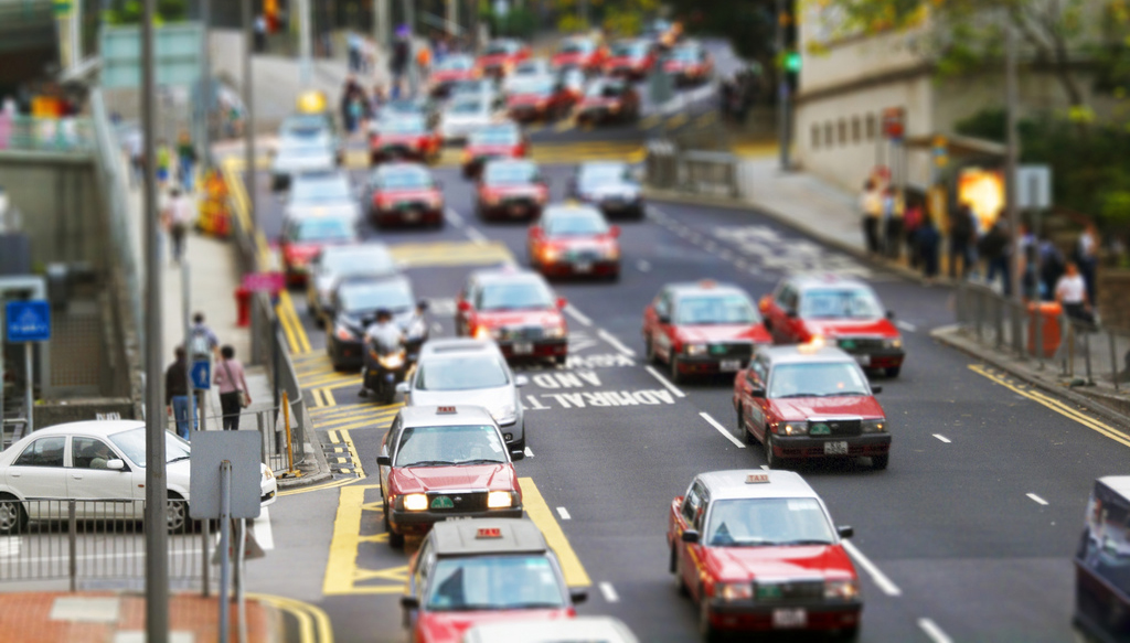 A tiltshift photo of a HK street bustling with red taxi cabs.