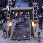 Banners hang on either side of a snow-covered Torii gate lit by lanterns.
