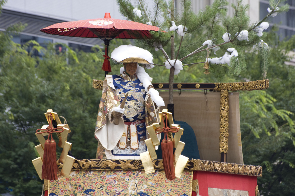 The crown of a float with an elderly female figure dressed in robes standing beneath a red parasol.