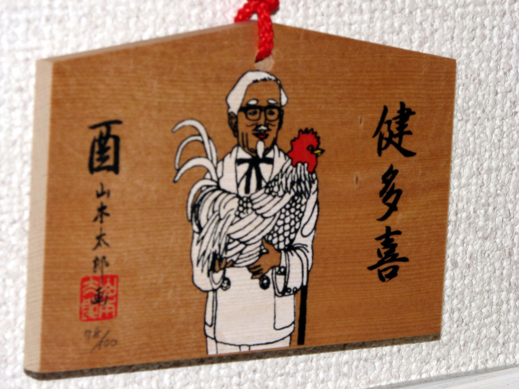 A small wooden prayer tablet painted with Colonel Sanders holding a rooster in his arms.