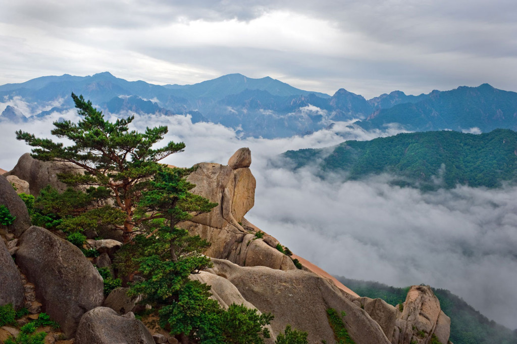 Clouds drift beneath green mountain peaks where a hanging stone teeters at the edge of a cliff.