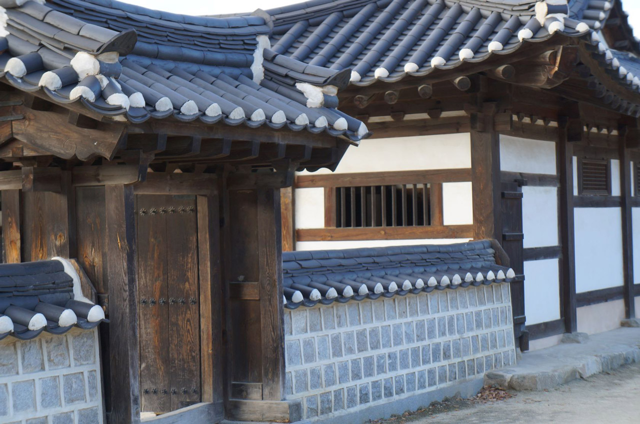 The front of a traditional korean home with a blue clay tile roof and white walls structured with heavy wooden framing.