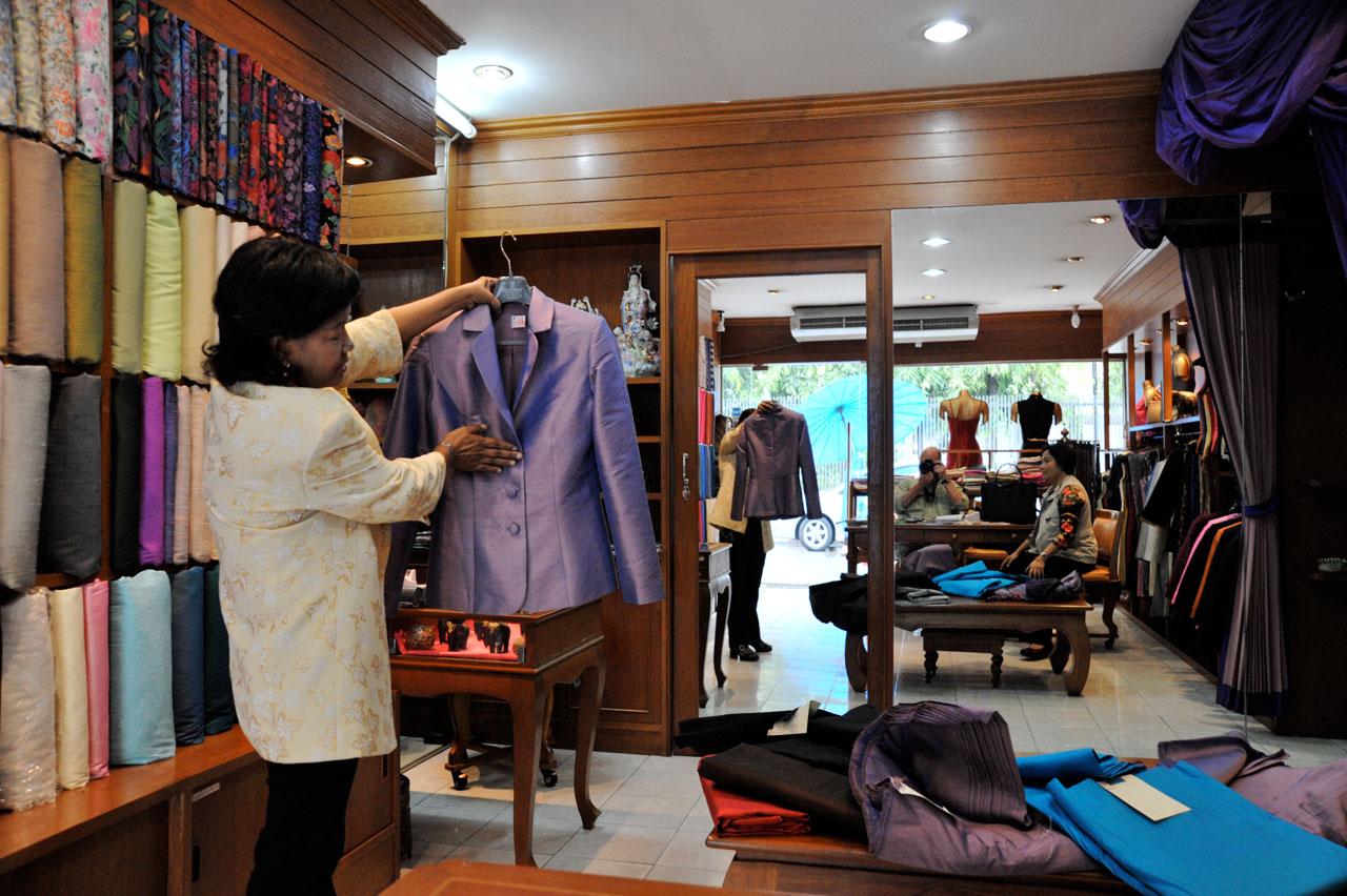 Bolts of cloth are shelved and a tailor examines a custom made jacket.