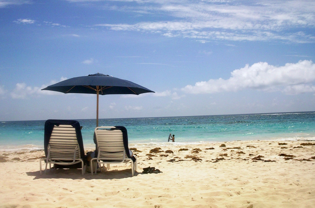 In pale golden sand, two beach chairs under a blue umbrella face the aqua water.