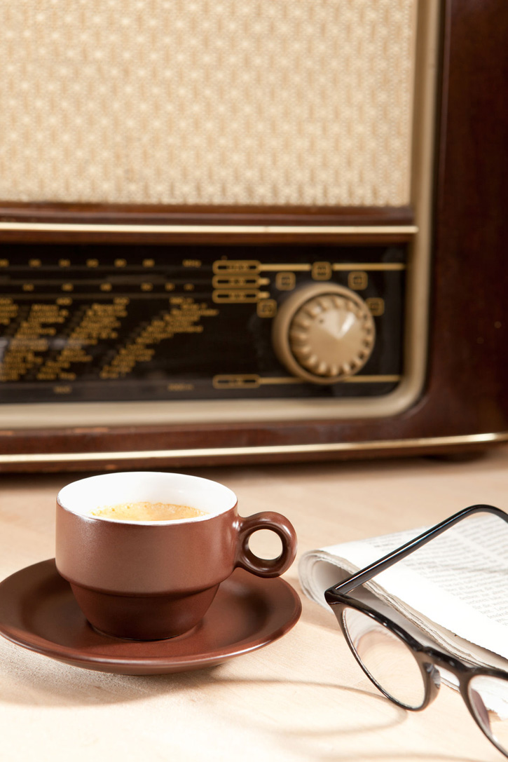 A cup of coffee in front of a vintage radio, newspaper and eyeglasses.