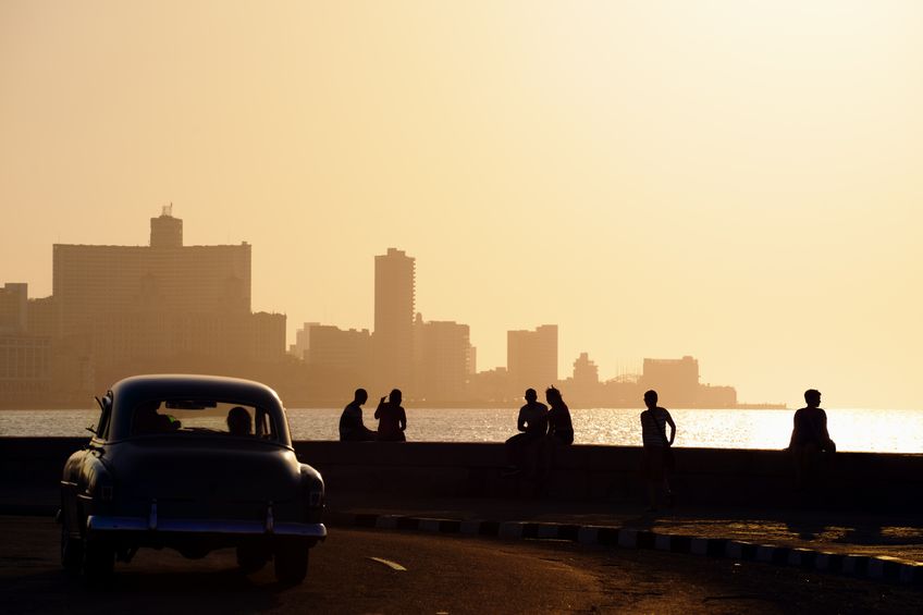 People sitting on the Malecon at sunset as vintage cars drive by in Havana, Cuba.