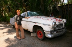 Ada Rosa stands beside a white rusting Chrysler parked on the dirt.