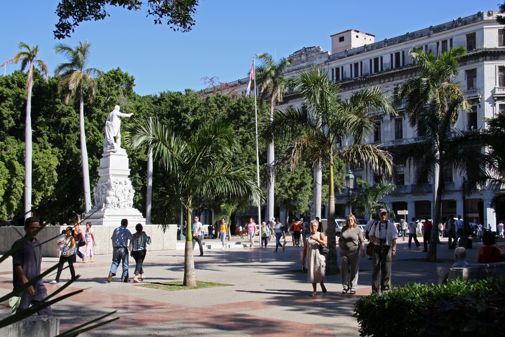 Pedestrians stroll through a plaza studded with palm trees and featuring a statue.