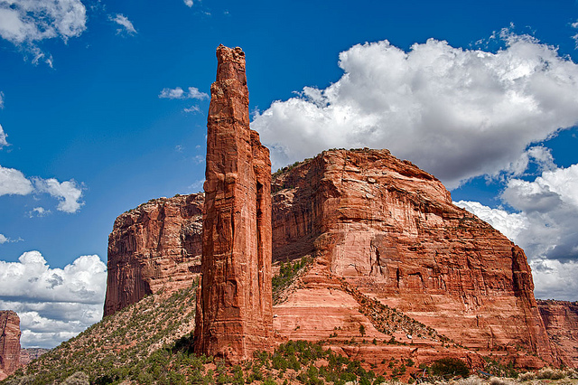 A tall pillar of red rock juts skyward from the floor of a deep canyon with a massive plateau of rock visible behind it.