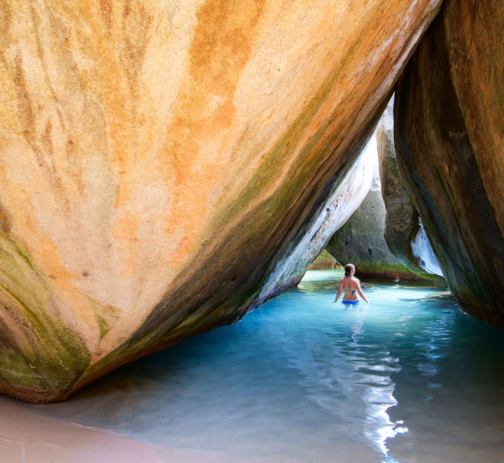 A woman wades in a pool formed by the intersection of two large rocks that allow in a small shaft of sunlight.