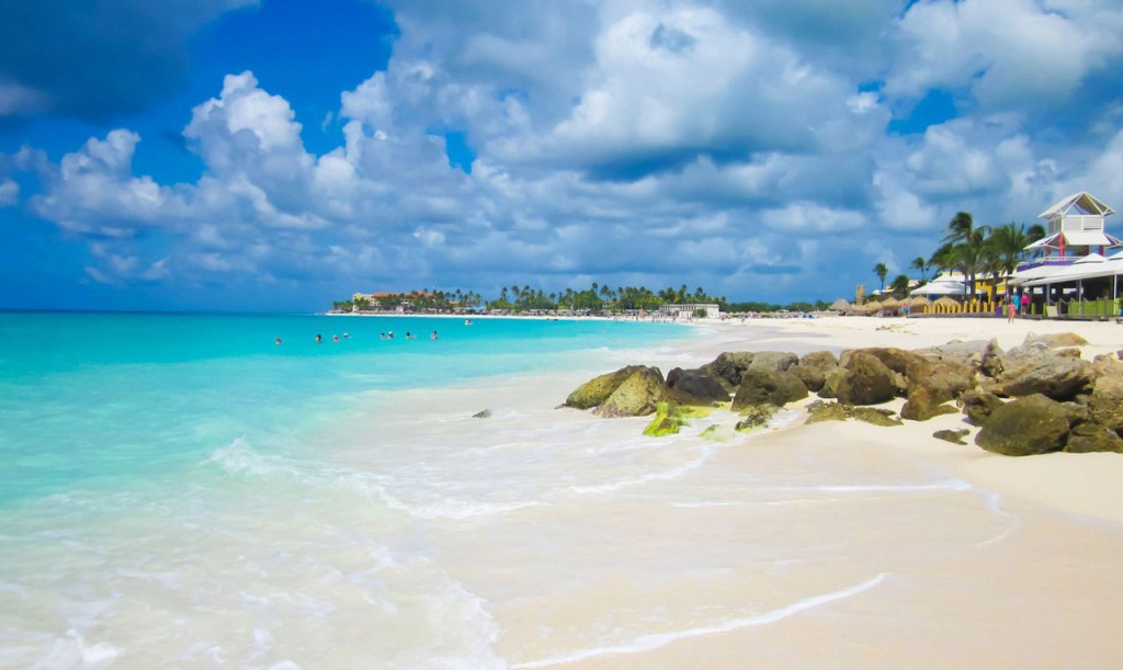 Soft waves lapping onto a wide white sand beach with swimmers in the water near a beachfront resort in the distance.