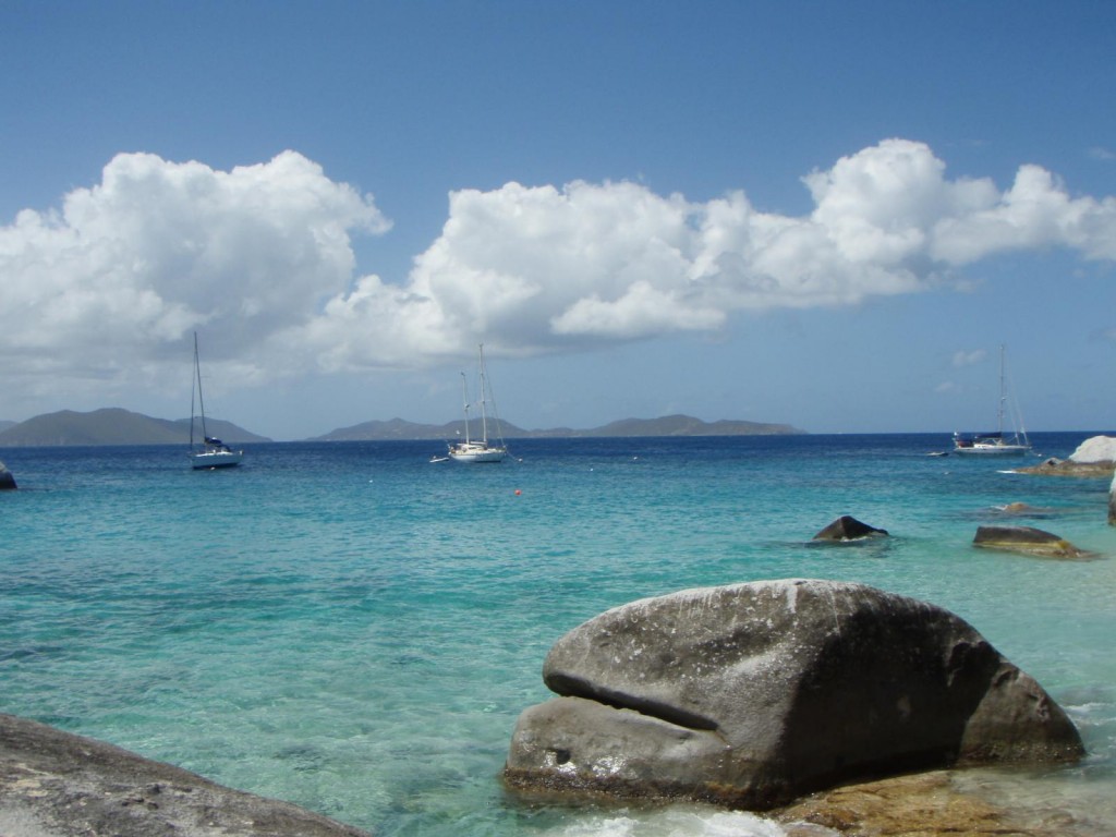 Large boulders in clear turqoise waters with sailboats and large puffy clouds in the distance.