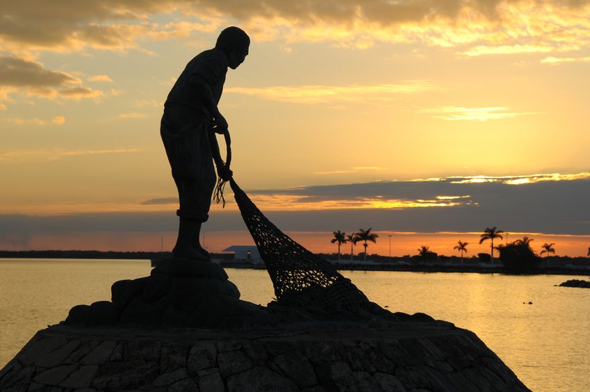 At sunset on the coast, the silhouetted statue of a fisherman hauling in his net.