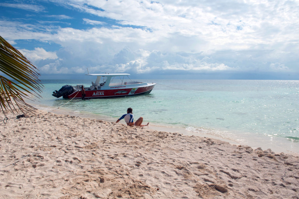 A person in a rash guard sits on the sand with a boat anchored just off-shore.