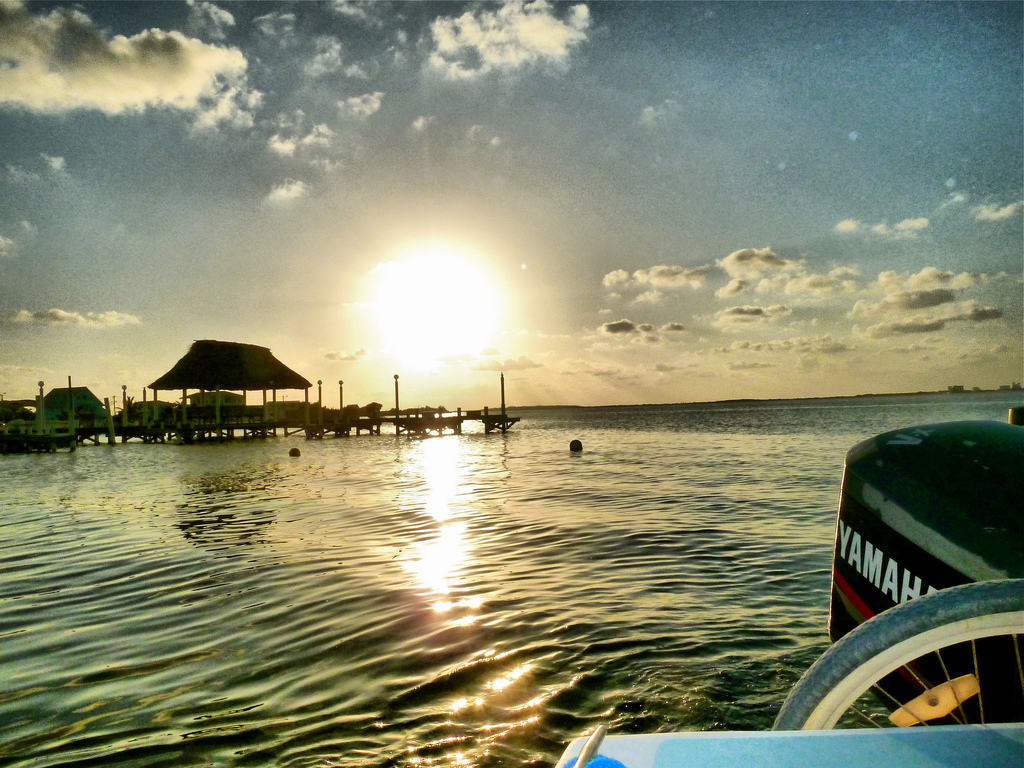 View from the back of a water taxi with the sun going down behind a dock extending into the water.