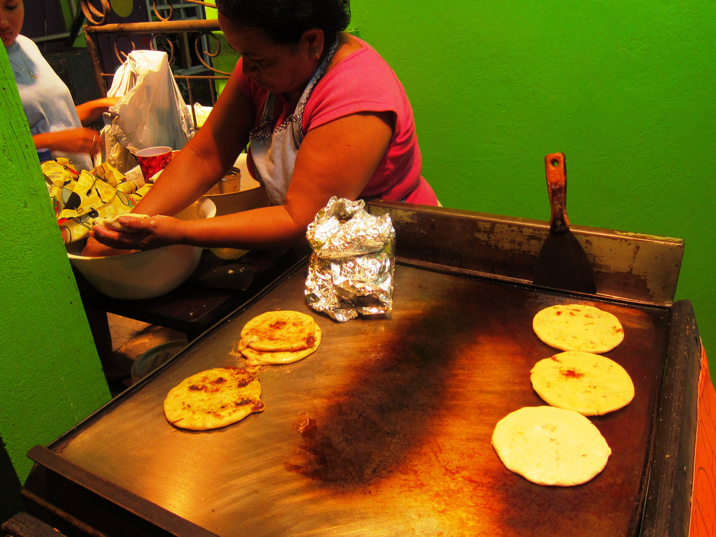 A woman forms pupusas by hand, cooking them on a griddle surface next to her.
