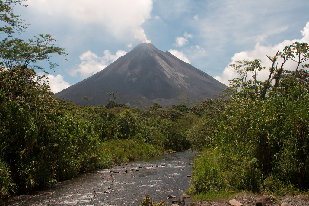 A small river runs in the foreground while the near perfect triangle silhouette of Mount Arenal rises in the distance.