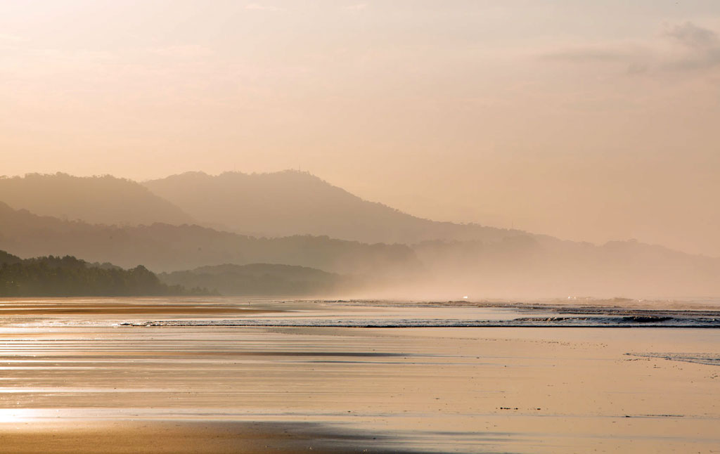 Hills recede in the distance as hazy orange light spills across a wide flat beach.