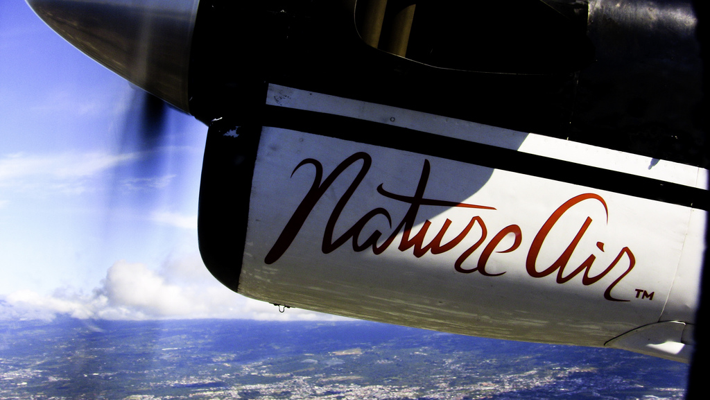 View out a plane window of the land below and NatureAir written on the propeller housing.
