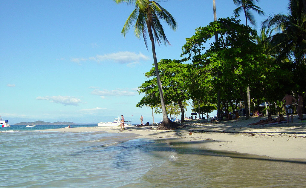 Shallow water surrounds a narrow golden beach studded with palm trees with a yacht visible in the distance.