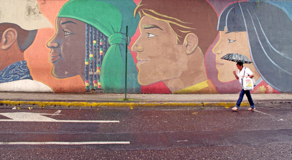 A woman walks with an umbrella past a street mural featuring multicultural portraits.