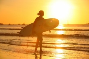 A surfer walks down the beach carrying her board with the setting sun behind her.