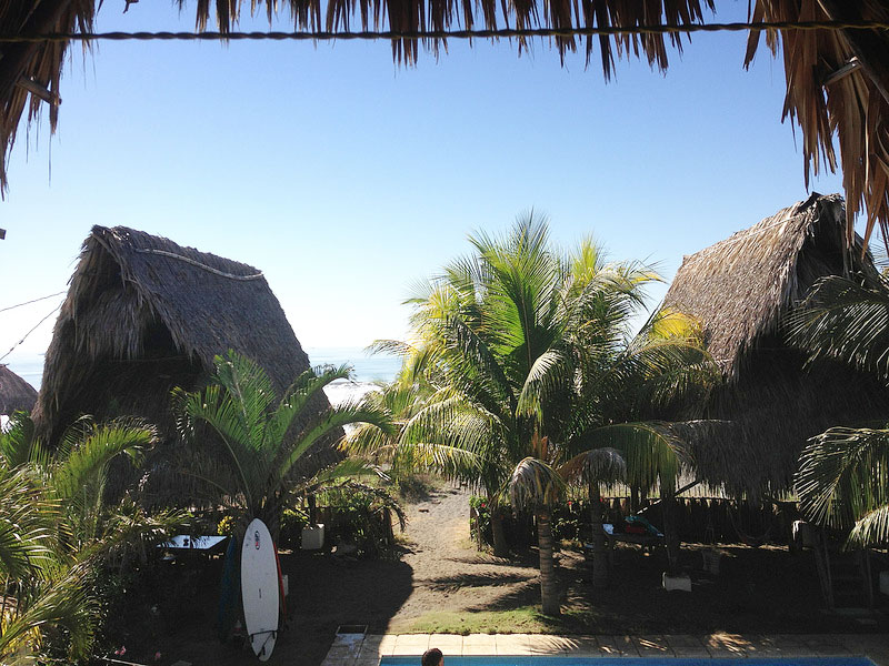 A surfboard sits propped against tables poolside near palapas on the shore.