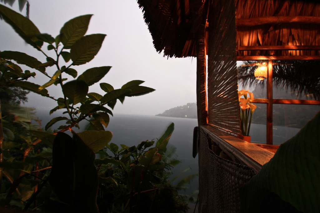 Warm lighting from a palapa-covered room contrasts with light fog on the nearby lake.