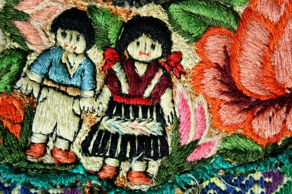 Colorful embroidered textile with two figures of kids holding hands amidst a floral pattern.