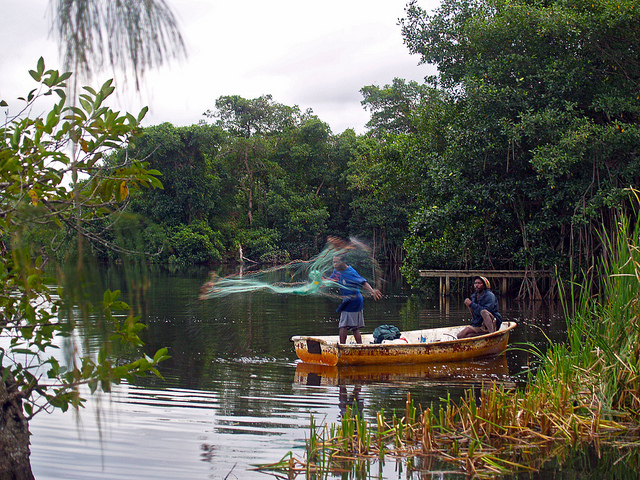 A pair of fisherman in a small rowboat cast a net into calm river water.