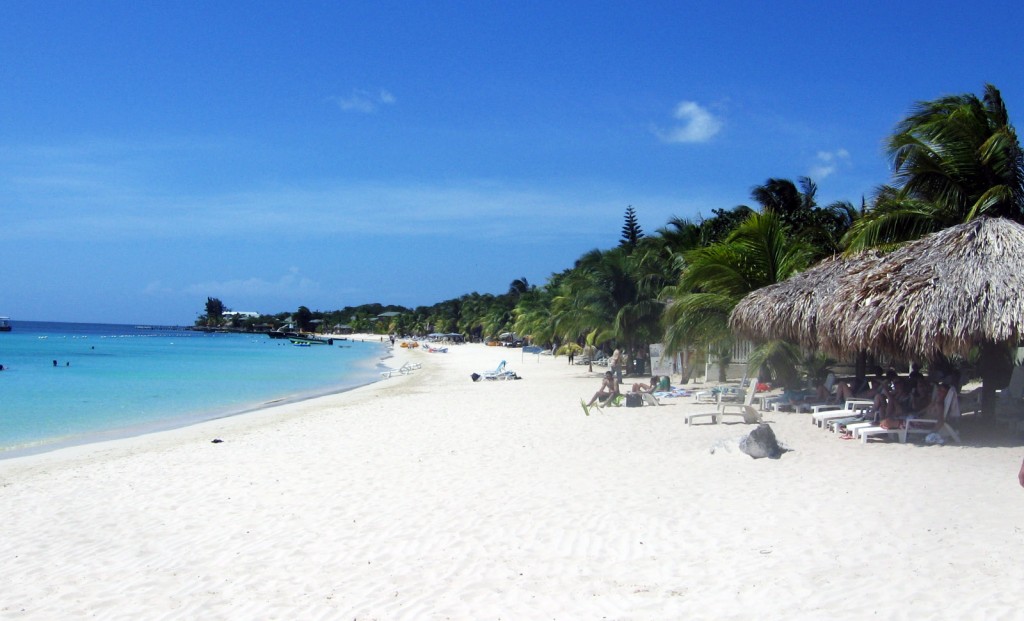 A white sand beach stretches into the distance with lounge chairs shaded by a thatched overhang.