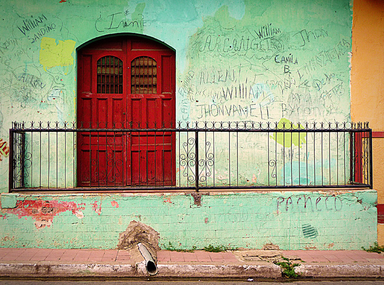 A red door in a pale green wall surrounded by graffitied names.