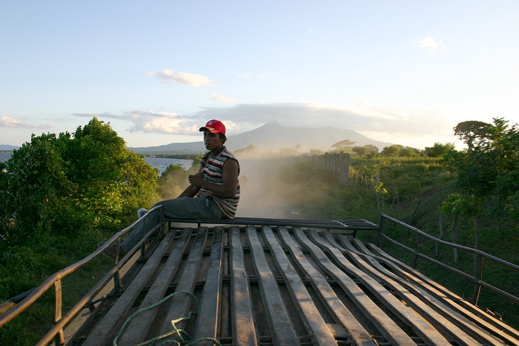 Dust rises as a man perches on the roof of a chicken bus with the view of the volcano in the distance.
