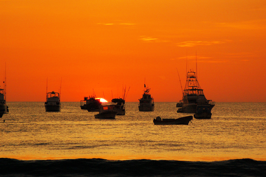 Fishing boats are silhouetted against a vibrant orange sky.