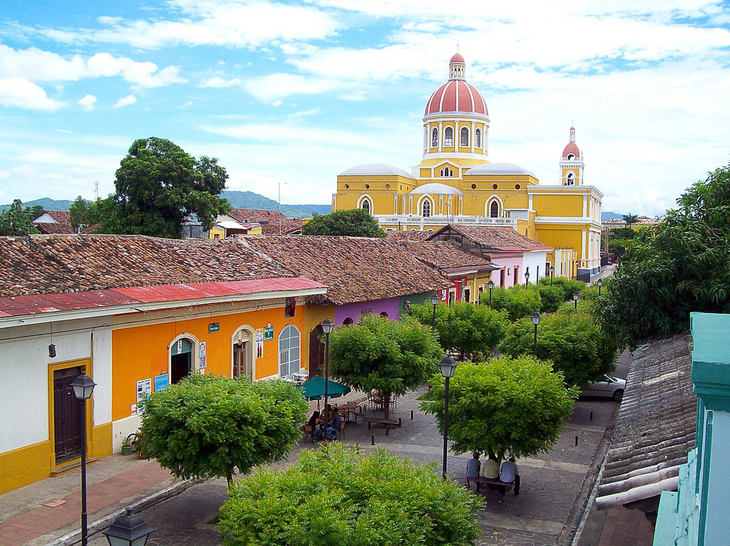 Trees dot a courtyard running between colorful buildings with a yellow, colonial style church rising a block away.