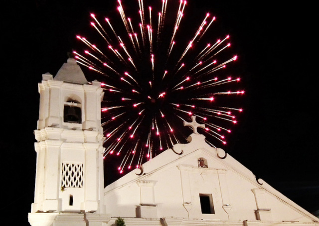 A red burst of fireworks goes off in the dark sky above the simple church.