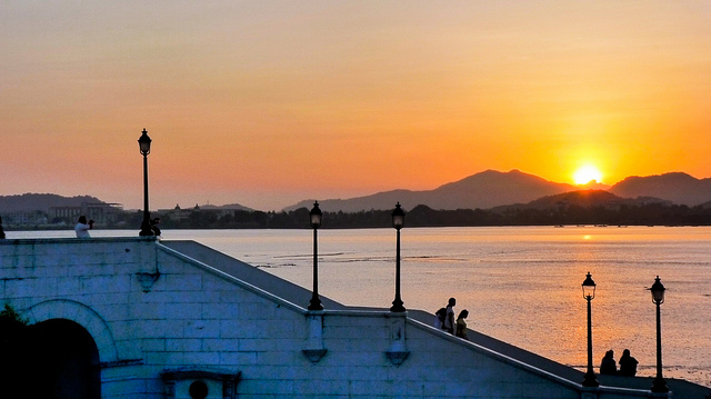 Pedestrians on a walkway with the sun disappearing over the horizon across the water.