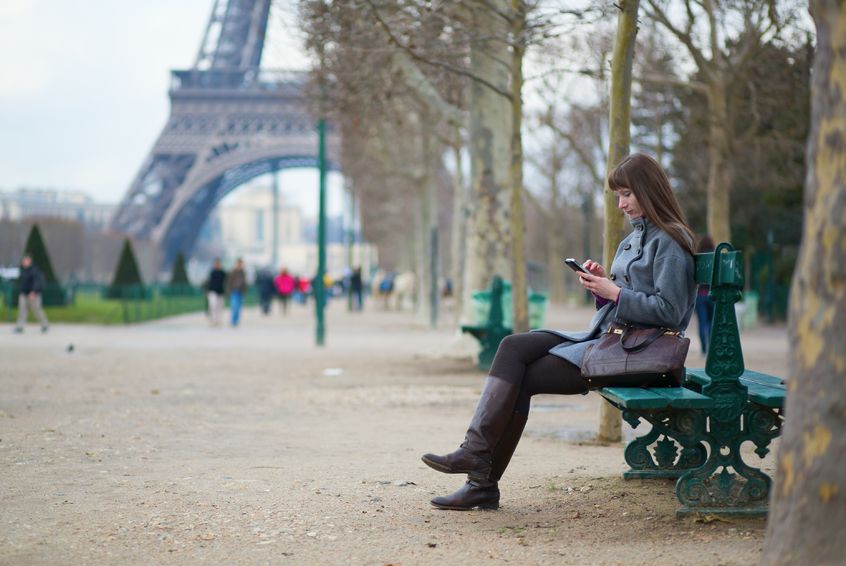 A woman at a park bench texts on her phone with the Eiffel Tower visible in the distance.