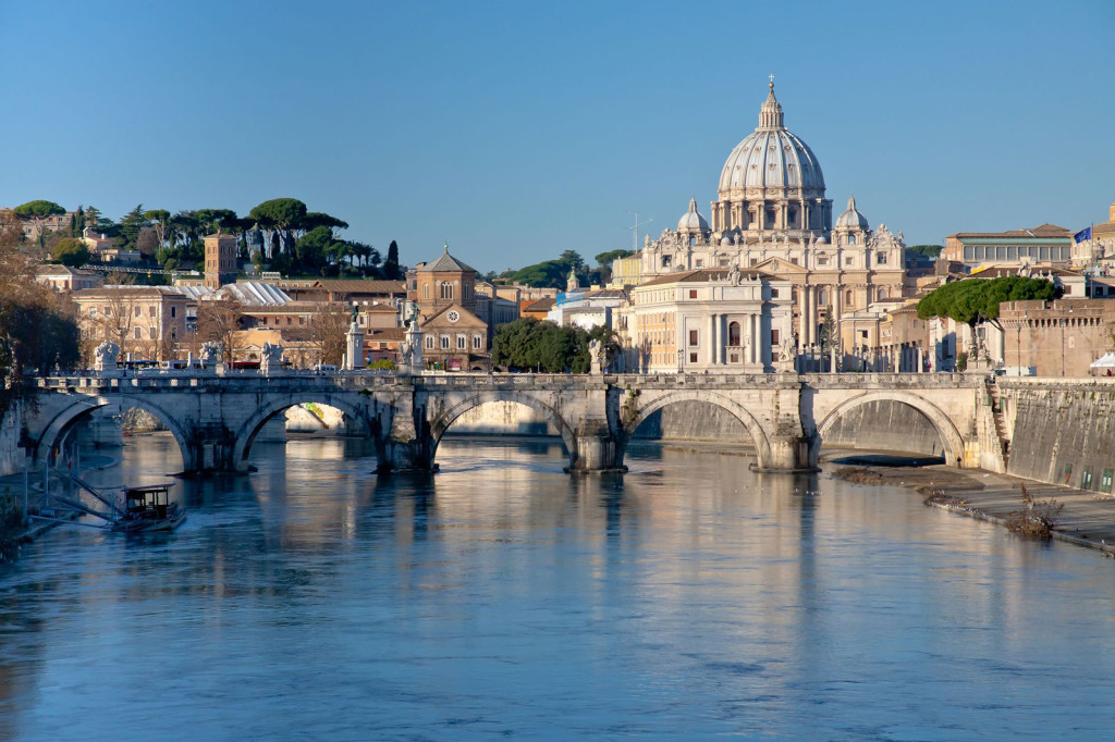 A stone bridge studded with sculptures crosses the Tiber with a view of the dome of St. Peter's Basilica rising just beyond.